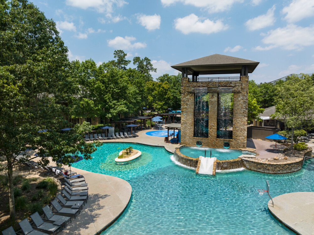 A large pool surrounded by lounge chairs and trees a tower with water falls and water slides in the middle.