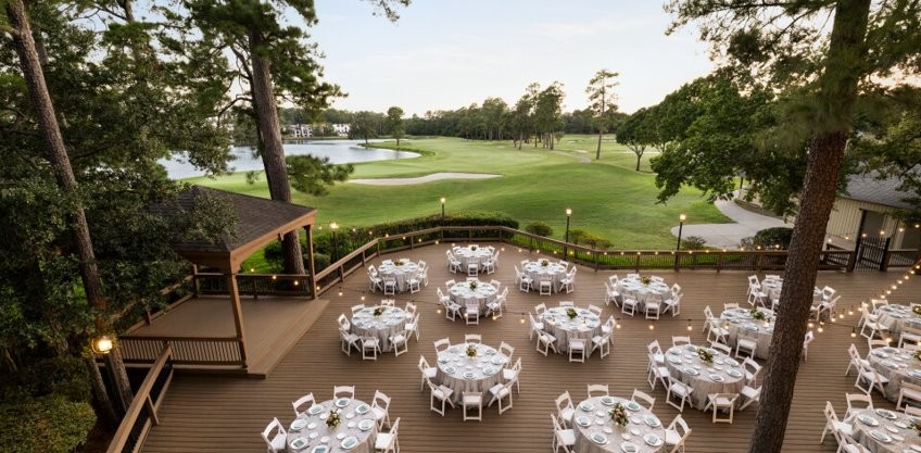 Outdoor event space on a deck overlooking the golf course. Round tables draped in white tablecloths and white folding chairs are spread out over the deck while while string lights hang above.