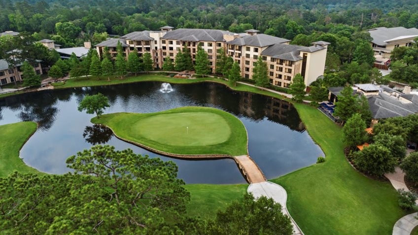 Aerial shot of the resort, with a golf green in the middle of a pond. Trees surround the course and buildings.